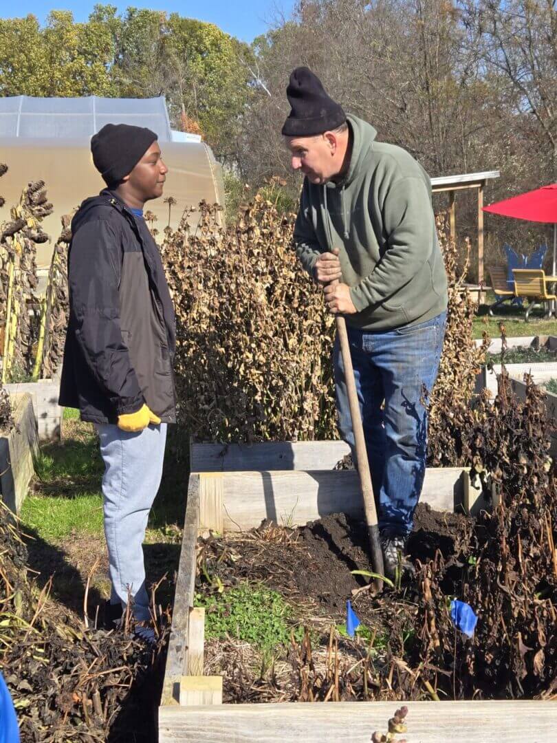 Two men conversing in a garden, one holding a shovel.